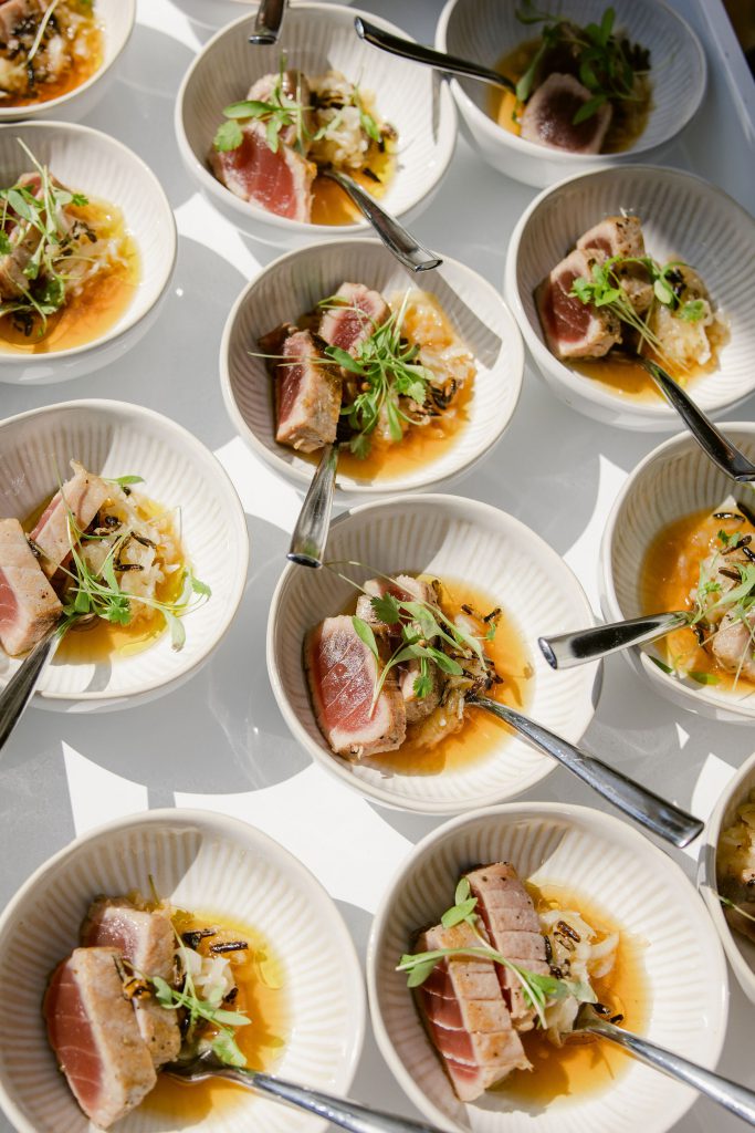 Individual bowls of seared tuna garnished with greens and a light sauce, each with a small fork, arranged in a grid pattern on a white surface.