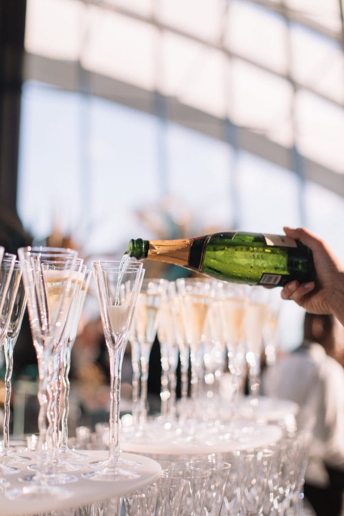 A large indoor event with people gathered under a high, curved glass ceiling, offering a panoramic view of a city skyline. Tables with chairs and plants are arranged around the space, creating a lively and elegant atmosphere.