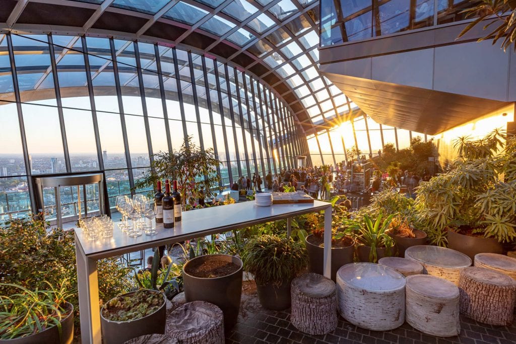 Rooftop garden bar with panoramic city views at sunset. Large glass windows, lush greenery, and drinks on tables create a vibrant atmosphere.