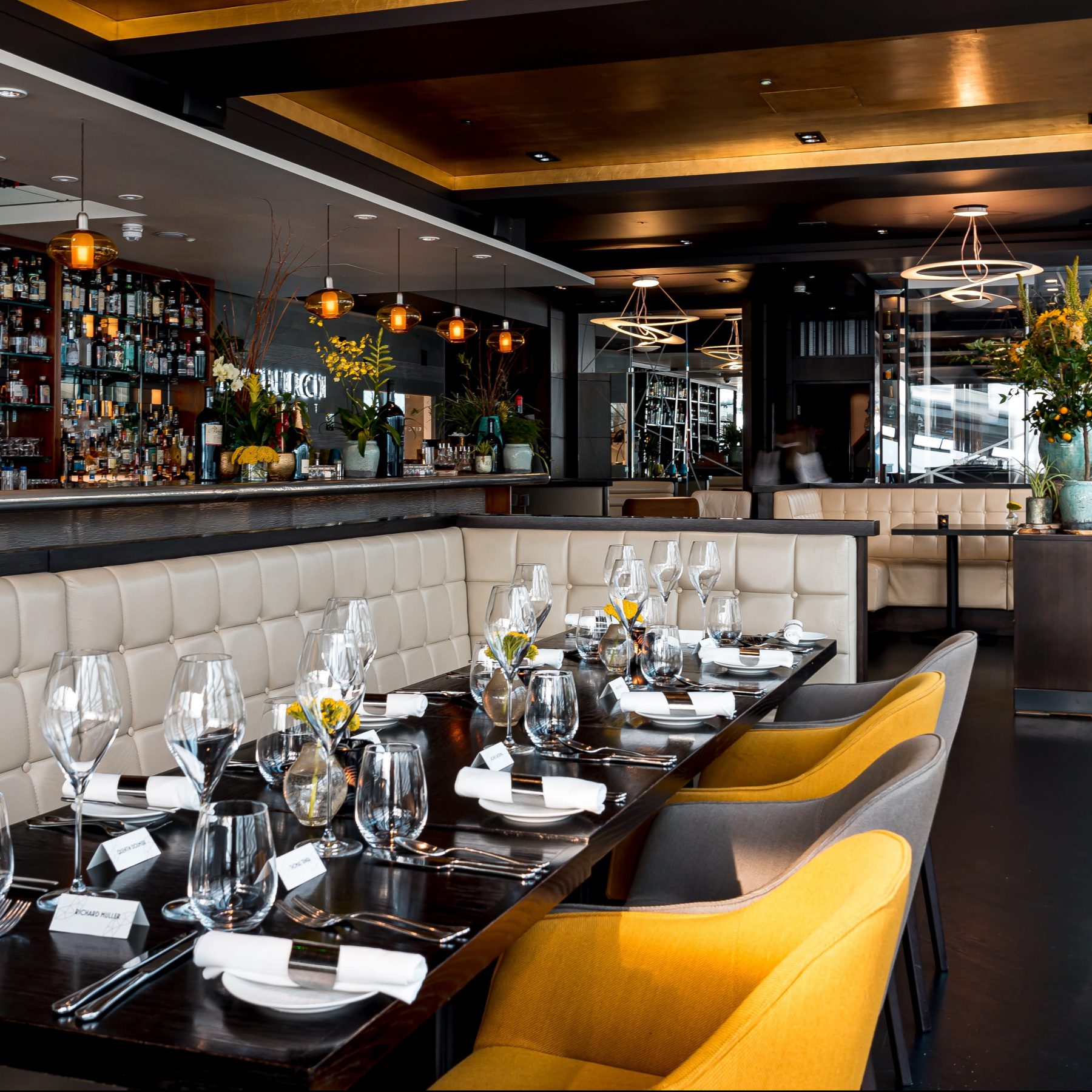 A modern restaurant interior with elegant table settings, featuring white cushioned booths and mustard yellow chairs. The bar is stocked with bottles, and pendant lights hang from the ceiling. Large windows provide ample natural light.