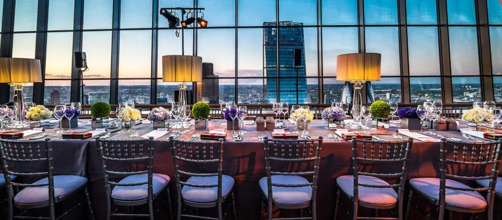 A long dining table set elegantly with plates, glasses, and floral centerpieces is positioned in front of a large window. The window offers a panoramic view of a cityscape at sunset, enhancing the ambiance with warm lighting.