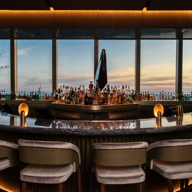 A sleek, modern bar with plush stools and a curved counter, lined with bottles and soft lighting. Large windows offer a panoramic sunset view over the ocean, enhancing the elegant ambiance.