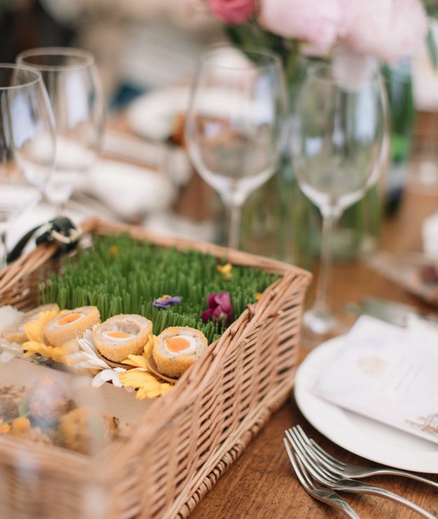 A bottle of white wine with a filled wine glass sits on a grassy field during sunset. Nearby are a basket, yellow flowers, and some bread rolls wrapped in paper, creating a picnic setting.