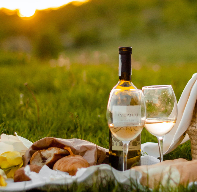 A bottle of white wine with a filled wine glass sits on a grassy field during sunset. Nearby are a basket, yellow flowers, and some bread rolls wrapped in paper, creating a picnic setting.