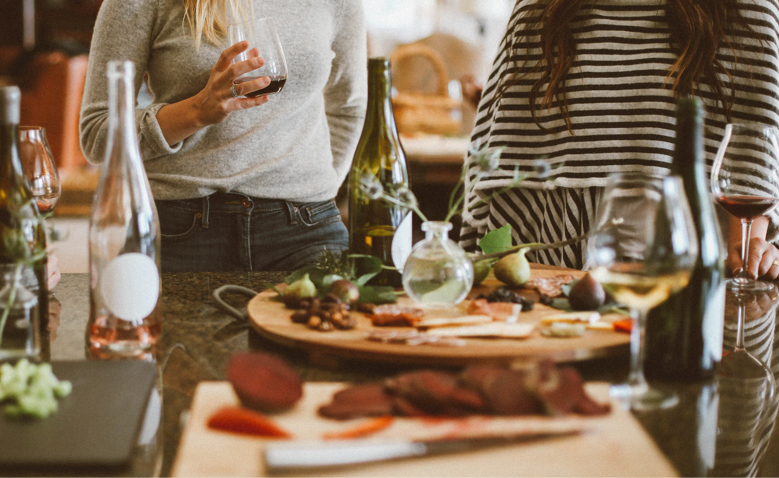 Two individuals stand at a counter with wine glasses. The table is set with wine bottles and platters of food, including sliced meats and vegetables. The scene is casual and relaxed, suggesting a social gathering or informal meal.