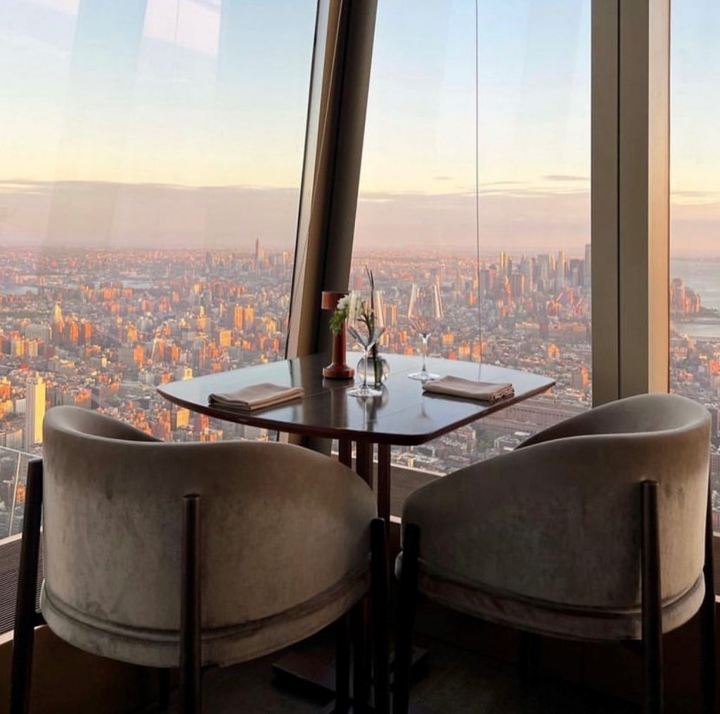 A dining table set for two with elegant chairs is positioned by large windows overlooking a sprawling cityscape at sunset. The skyline is illuminated with warm golden hues, creating a romantic and scenic atmosphere.