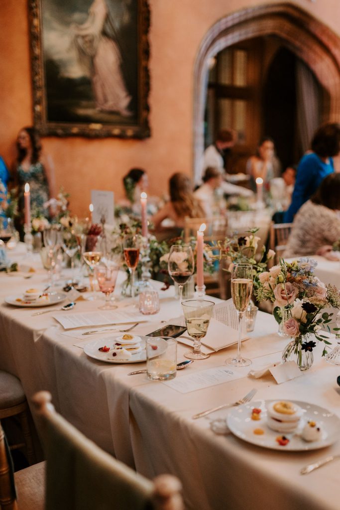 A beautifully set dining table at an elegant event, adorned with floral centerpieces, candles, and various plated dishes. Guests are seated and conversing in the background, with an ornate painting and archway visible on the walls.