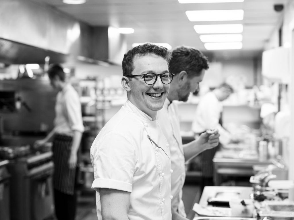 A smiling chef wearing glasses and a white uniform stands in a bustling kitchen, with several other chefs working in the background. The setting is lively and well-lit, conveying an atmosphere of teamwork and culinary activity.