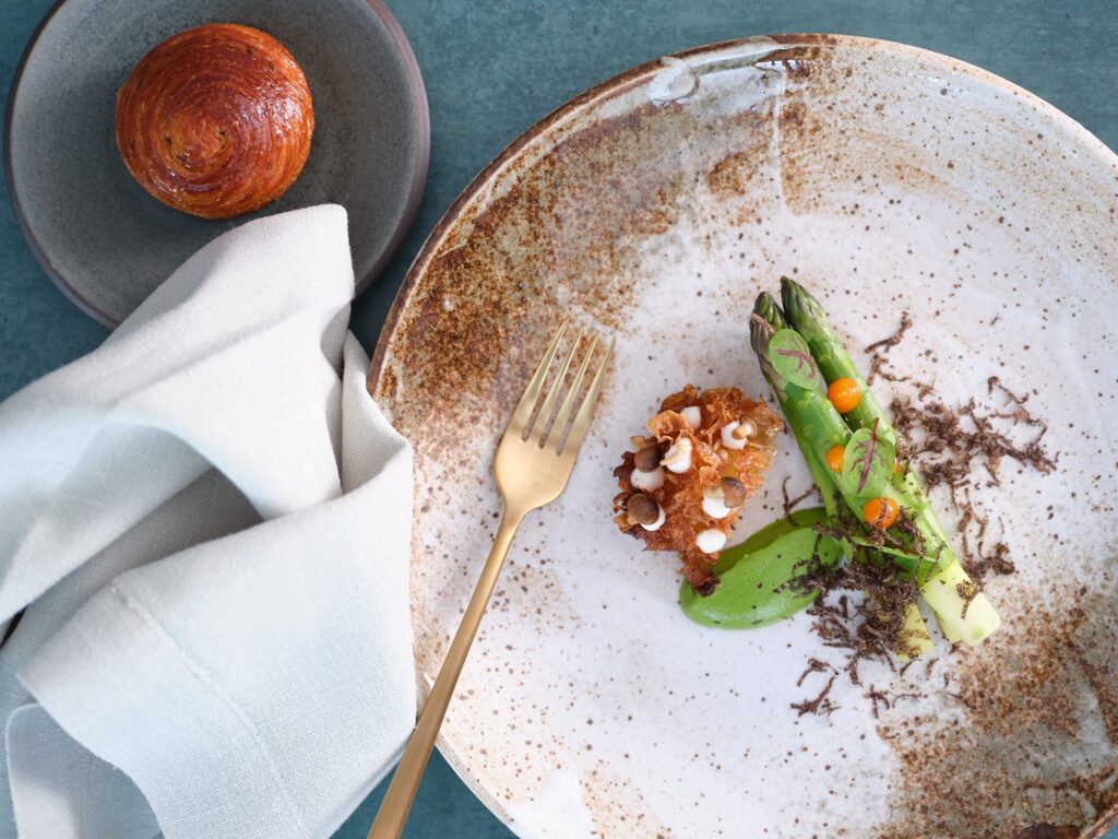 A gourmet dish featuring asparagus, a green sauce, and a textured garnish served on a large, rustic plate. A gold fork rests on the plate. A small round bread roll is on a separate dish nearby, next to a white cloth napkin.