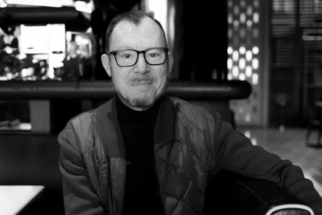 A black and white photo of a man with glasses, a short beard, and short hair, sitting in a restaurant. He's wearing a quilted jacket over a turtleneck sweater and smiling at the camera. The background shows blurred restaurant decor.