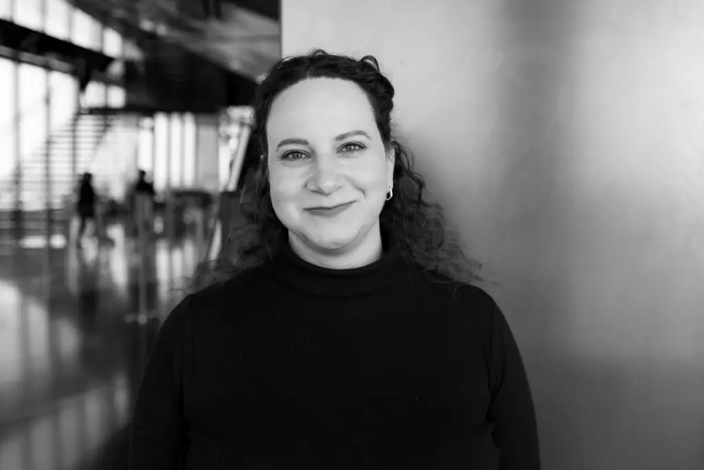 A person with curly hair and a smile stands indoors against a wall. The background features a modern architectural space with large windows and a staircase. The photo is in black and white.