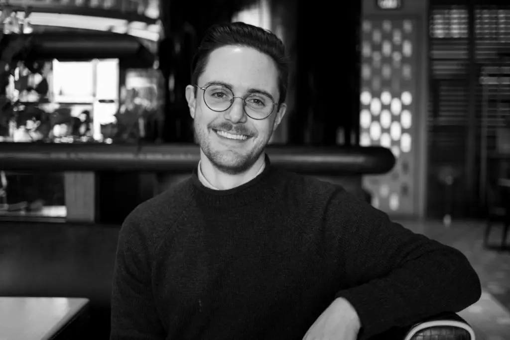 A person with glasses and a sweater smiles while sitting in a cozy, dimly lit cafe. The background features patterned walls and a soft, ambient lighting.