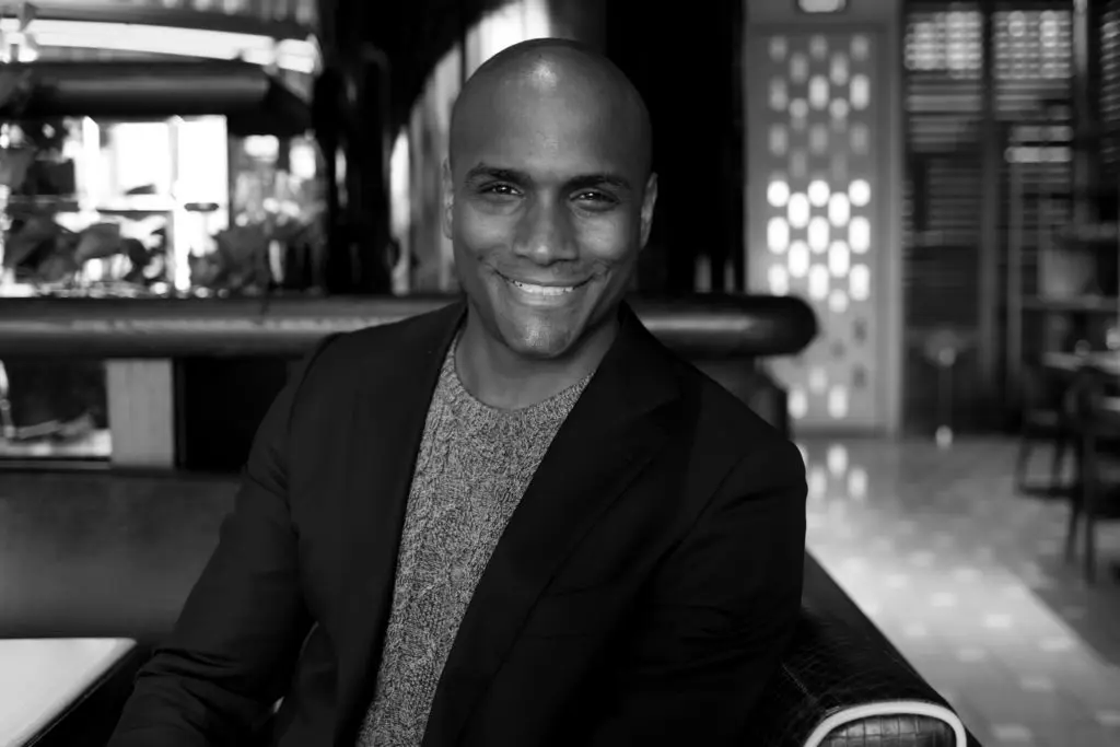 A smiling person with a shaved head is sitting in a stylish indoor setting. They are wearing a blazer over a sweater, and soft natural light is illuminating the space, creating a warm atmosphere.