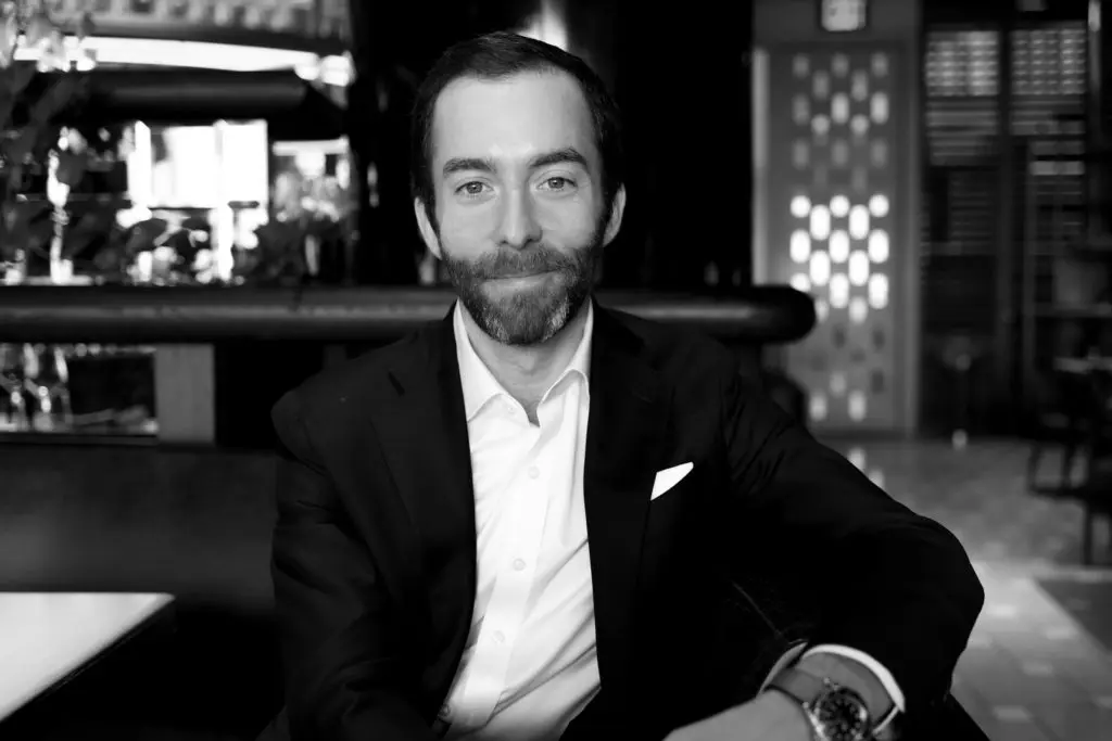 A man with a beard and wearing a suit sits confidently in a dimly lit room. The setting appears to be a stylish interior with a patterned background and soft lighting.