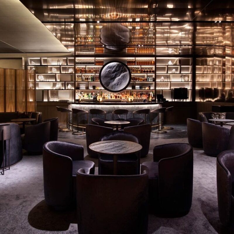 A sophisticated bar with a sleek design features dark plush seating, round tables, and a well-stocked backlit bar. A large circular mirror is mounted behind the bar, reflecting the luxurious ambiance of the dimly lit space.
