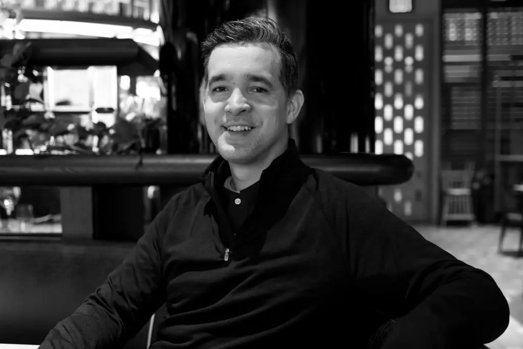 A person sitting at a table in an indoor setting, smiling at the camera. They are wearing a dark sweater and the background features geometric patterns on the walls with soft lighting. The image is in black and white.