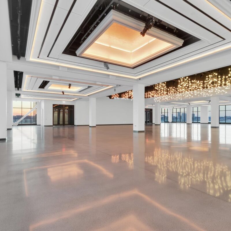 A spacious, empty event hall with polished floors and large windows offering a view of the city skyline. The ceiling features modern decorative lighting, including geometric panels and a cluster of small hanging lights. A sunset is visible outside.