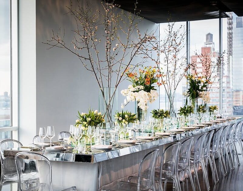 Long dining table set with elegant glassware and floral centerpieces, positioned by large windows overlooking a cityscape. Transparent chairs line the table, and tall branches with delicate blossoms provide a sophisticated and modern ambiance.
