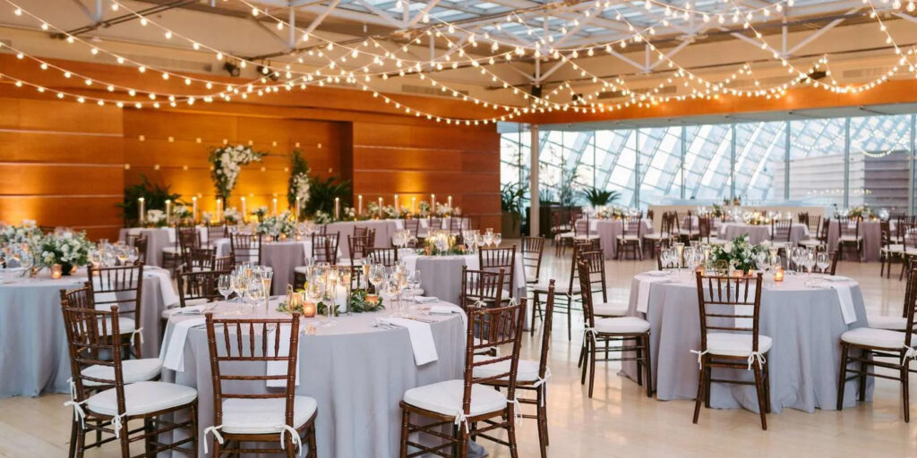 Spacious wedding venue with round tables covered in gray tablecloths, surrounded by wooden chairs. Tables are adorned with floral centerpieces and candles. Warm lighting and string lights create an elegant atmosphere. Large windows in the background.