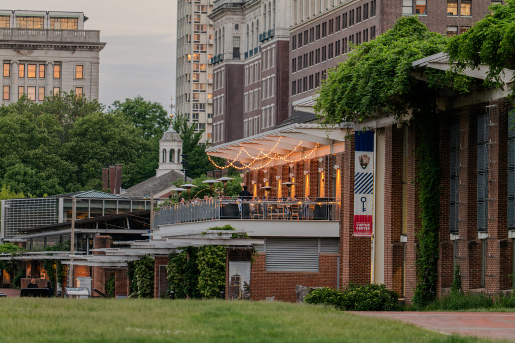 A scenic view of the cityscape highlights THE LIBERTY VIEW, where brick buildings are adorned with green ivy. String lights twinkle on a balcony offering outdoor seating. A small tower stands in the background, while Rhubarb Hospitality Collection caters to elegant events in Philadelphia's lush setting.