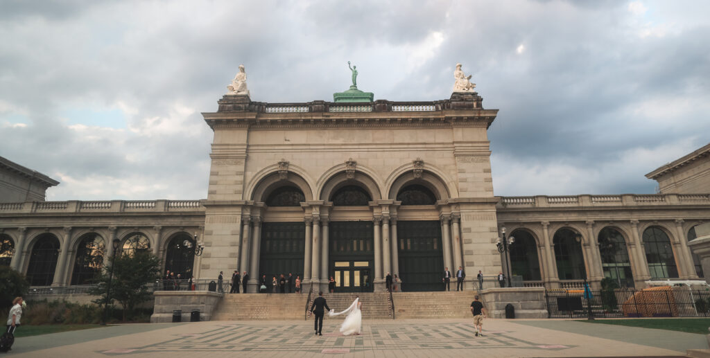 A couple stands on a large, open plaza in front of the grand Memorial Hall c. 1876, part of the Rhubarb Hospitality Collection in Philadelphia. Tall arches and statues grace the historic building, with a cloudy sky adding a dramatic backdrop to this captivating scene.