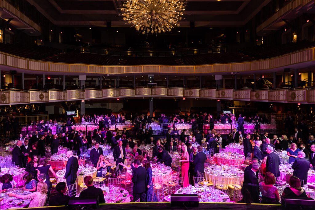 A large, elegant ballroom at THE MET PHILADELPHIA buzzes with guests in formal attire seated at round tables adorned with pink-lit centerpieces. A grand chandelier hangs overhead as the Rhubarb Hospitality Collection caters to the event, creating a vibrant atmosphere of mingling and conversation.