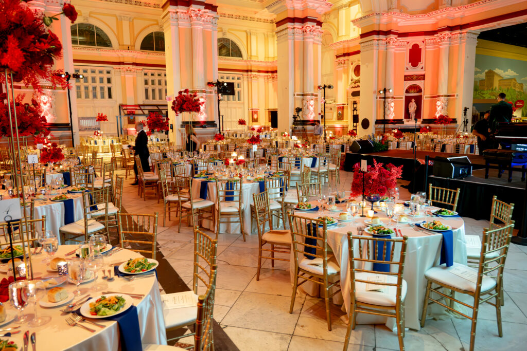 A lavishly decorated event hall features round tables with white tablecloths, gold chairs, and elegant table settings. Red floral centerpieces adorn the tables. The room is illuminated with warm lighting, highlighting the tall, ornate columns.
