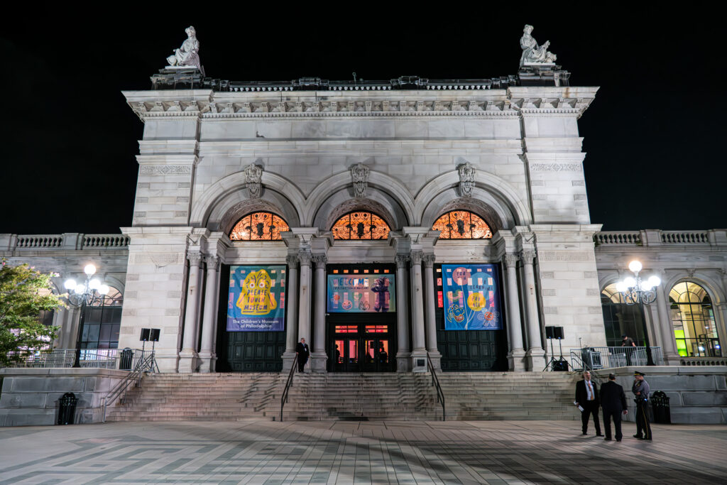 A grand, illuminated neoclassical building with three large arched entrances. Statues adorn the roof, and colorful banners hang above the doors. Several people stand on the steps leading up to the entrance.