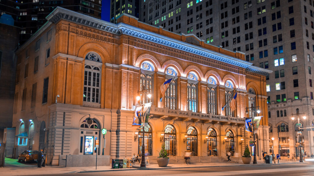 A large, illuminated historic building with arched windows and ornate architecture stands surrounded by tall skyscrapers in a city at night. Banners hang from the building, and streetlights illuminate the area. Photo Credit - G Widman.