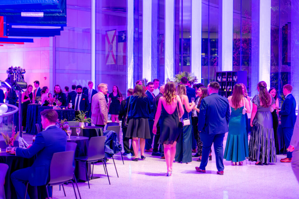 A vibrant event in a modern venue with large glass windows. People in formal attire, including suits and evening gowns, are socializing. Tables with drinks and floral arrangements are scattered around the room, illuminated by pink and purple lighting.