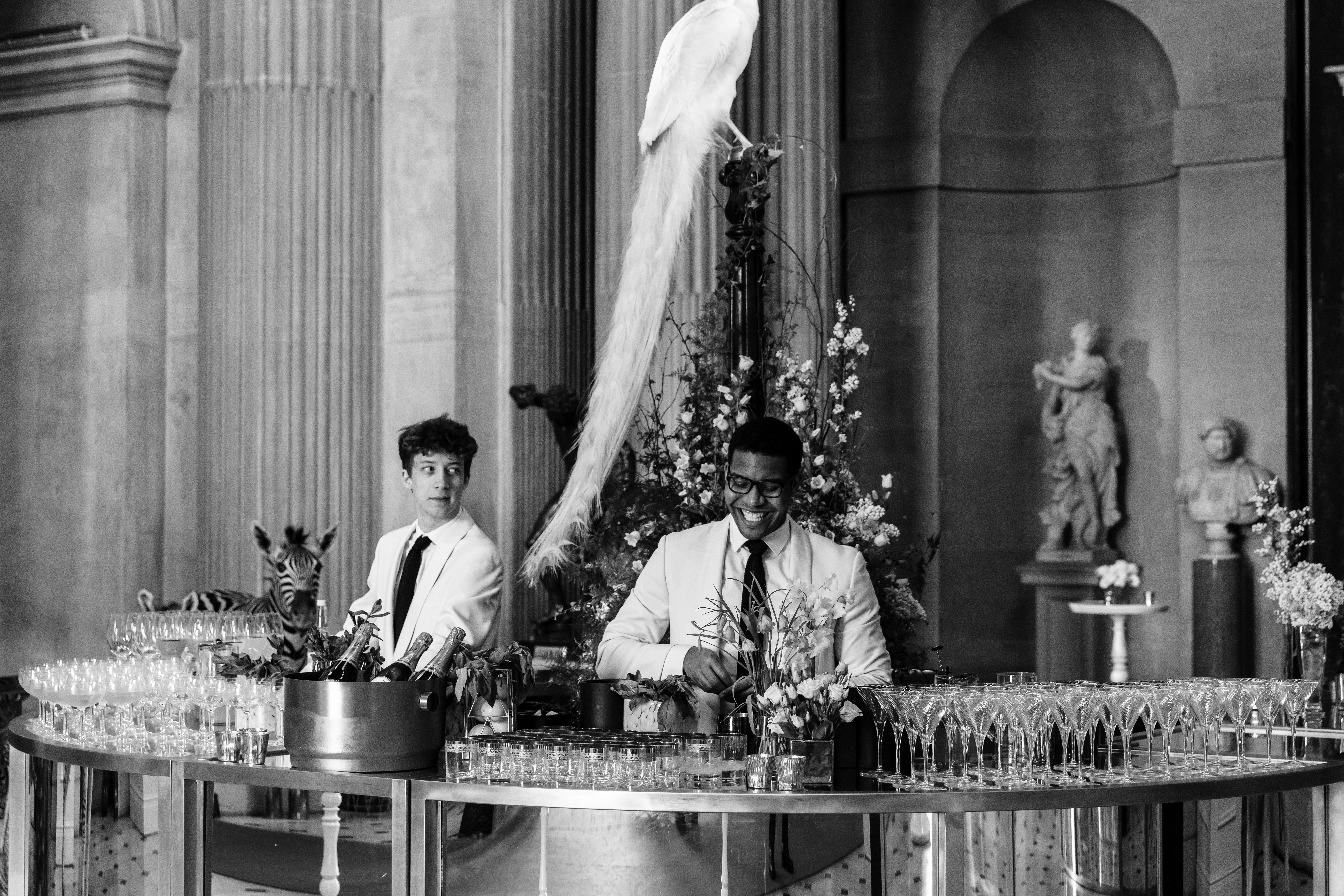 Two bartenders in white suits prepare drinks at an elegant venue with tall columns. A peacock decoration stands nearby, surrounded by flowers and numerous champagne glasses. Statue pedestals are visible in the background. Black and white image.