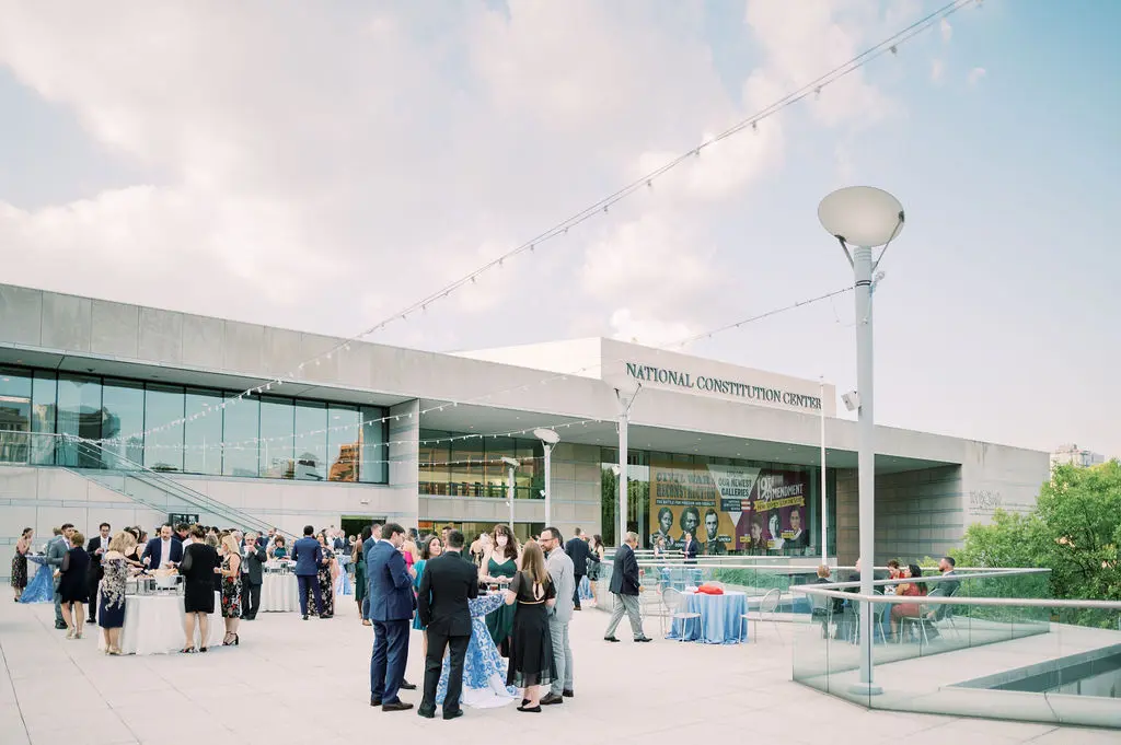 A group of people dressed in formal attire gather on a modern, open-air terrace with the National Constitution Center building in the background. String lights are overhead, and there are tables with white tablecloths around the space.