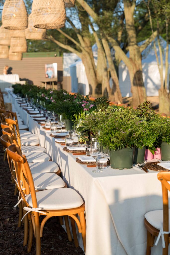 A long outdoor dining table with a white tablecloth, wooden chairs, and potted green plants as centerpieces is perfect for planning your dream wedding, set under hanging wicker lanterns among trees with neatly arranged place settings.
