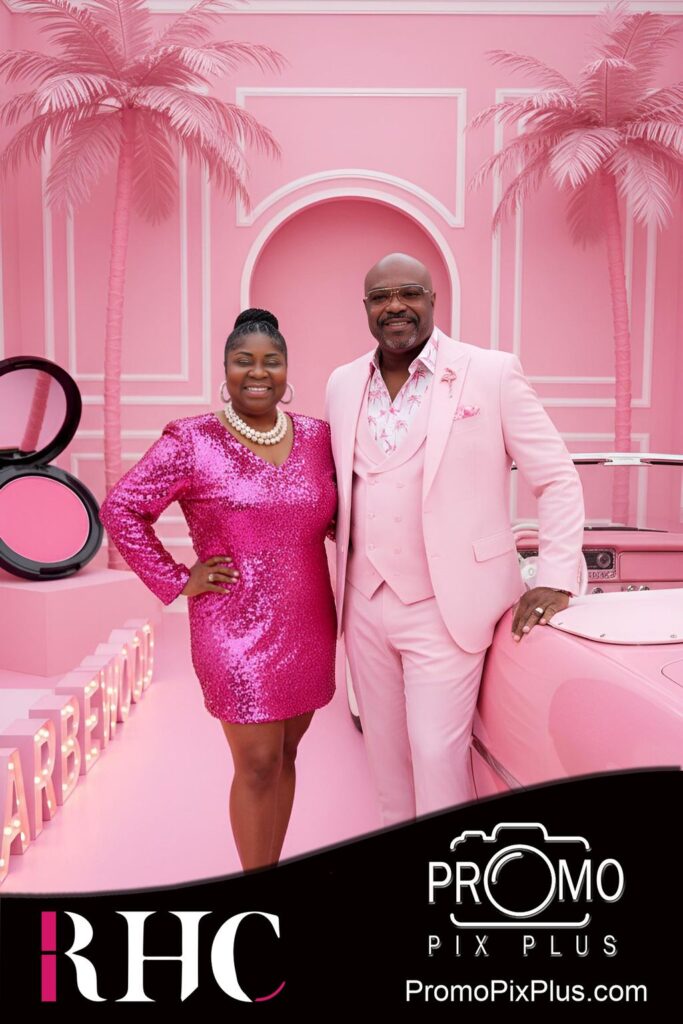 A smiling woman in a sparkly pink dress and a man in a light pink suit stand together in front of a pink car and a vibrant pink backdrop with “Barbiewood” lettering and palm tree designs.