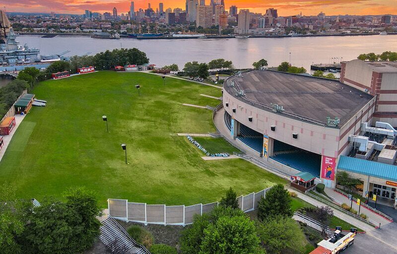 Aerial view of an outdoor amphitheater with a large grassy lawn facing a stage, set beside a river. At sunset, the city skyline and a docked navy ship are visible—reminiscent of venues like The Fillmore Philadelphia.